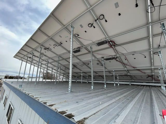 Solar panels installed on a metal roof, supported by metal frames and legs. Cables and wiring visible.