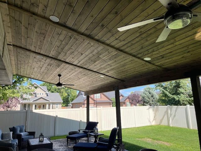 Covered patio with wooden ceiling, fans, and seating overlooking a yard with a white fence.