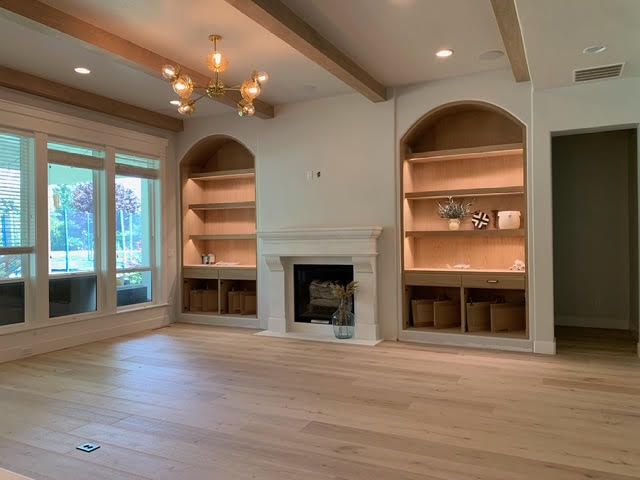 Empty living room with fireplace, built-in shelves, wood beams, light wood floors, and large windows.