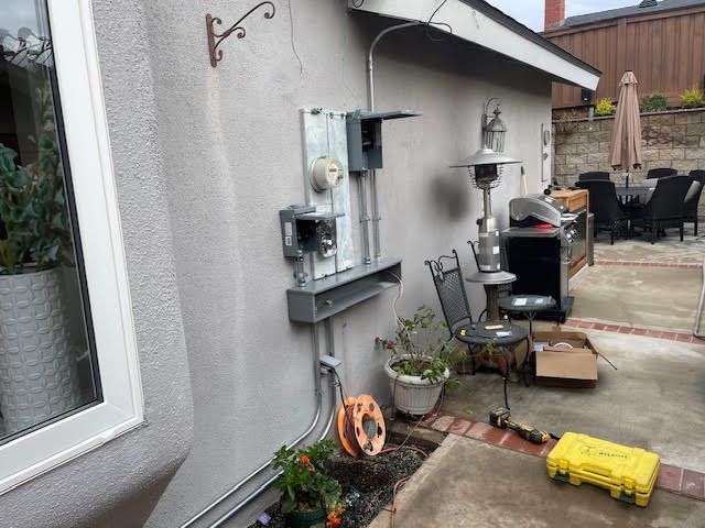 Electrical boxes and conduit mounted on a stucco wall outside, next to a window and patio.