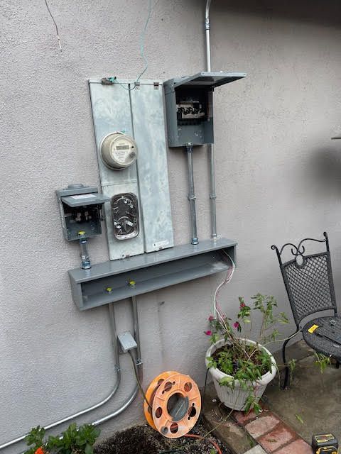Electrical meter and boxes mounted on a stucco wall outside. Gray metal components, orange wire spool, potted plants.