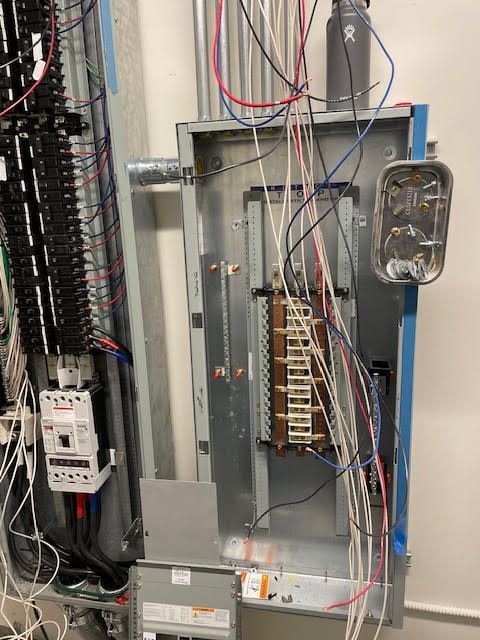 Electrical panel with wires, conduits, and circuit breakers against a white wall.
