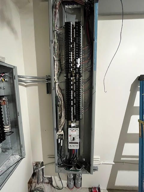 Open electrical panel with wires and circuit breakers. Gray metal box on white wall.