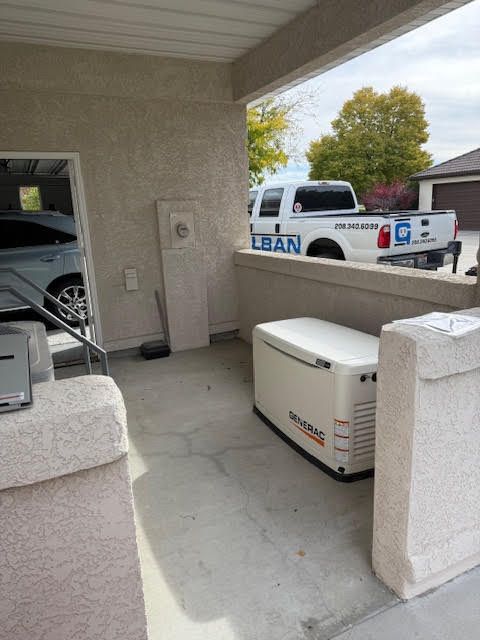 A white generator sits under a covered area next to an electrical panel. A truck with a logo is visible in the background.