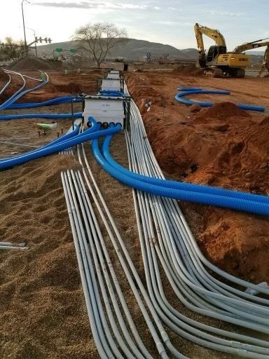 Construction site with buried blue and white conduit, connecting to utility boxes; dirt and heavy machinery.