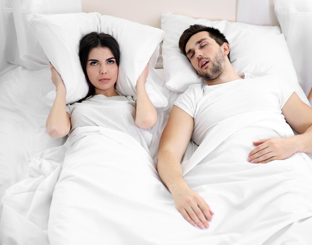 A man is snoring while a woman covers her ears with pillows.