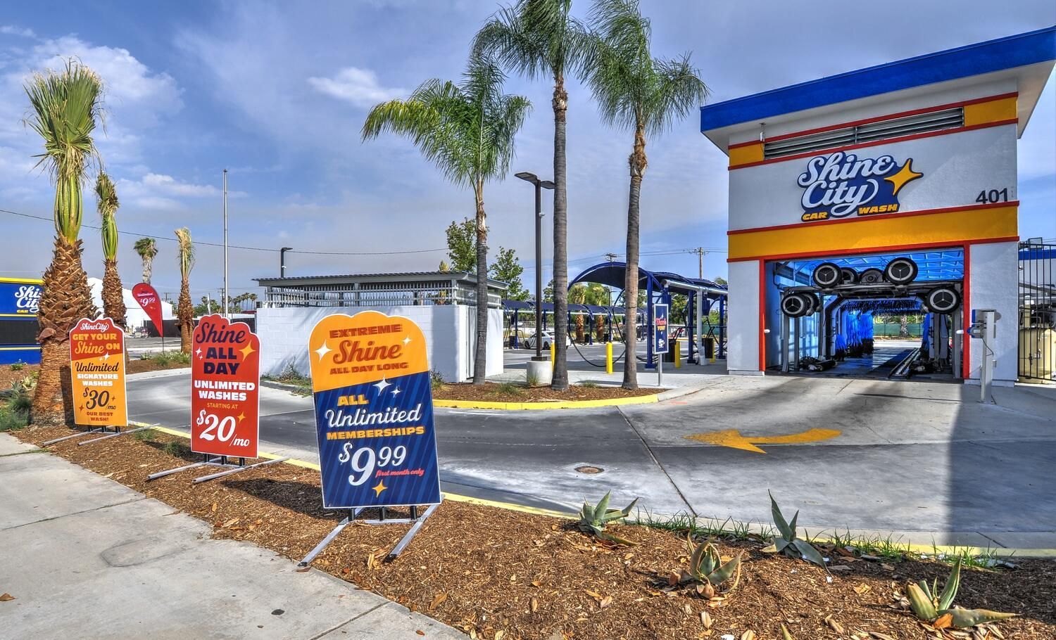 Exterior view of a car wash with signs advertising services, palm trees, and blue sky.