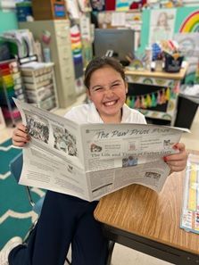 A young girl is sitting at a desk reading a newspaper.