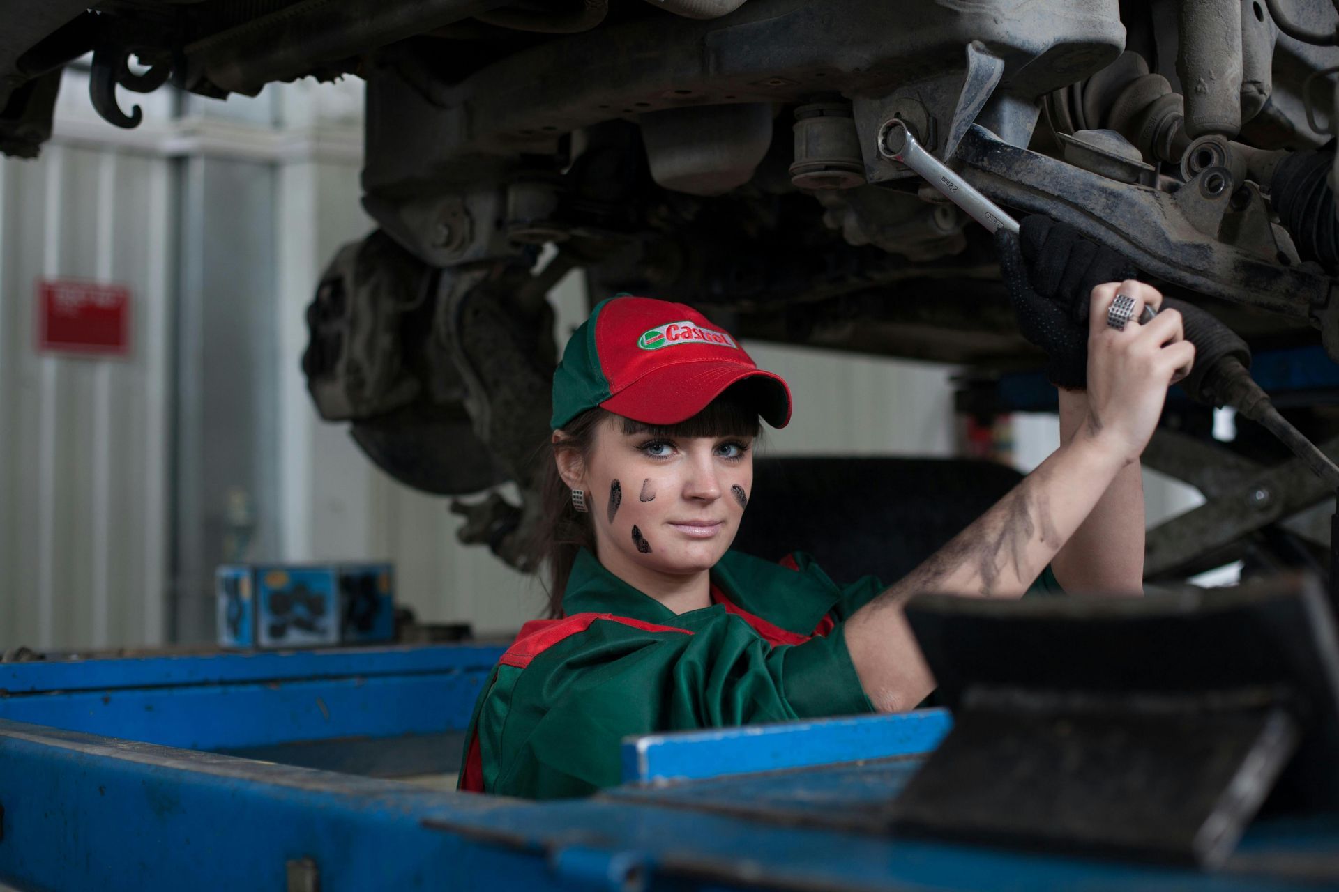 Woman mechanic under car, smiling, wearing hat, green uniform, face and arms smeared with grease.