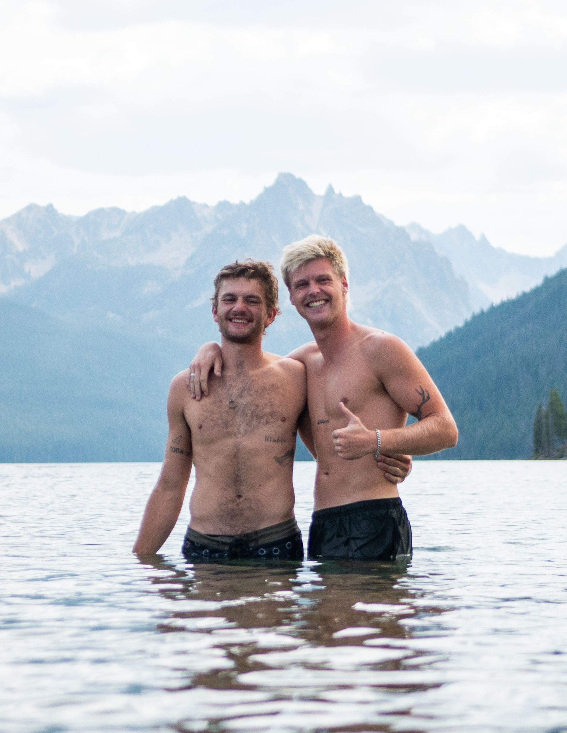 Two shirtless men are standing in a lake with mountains in the background.