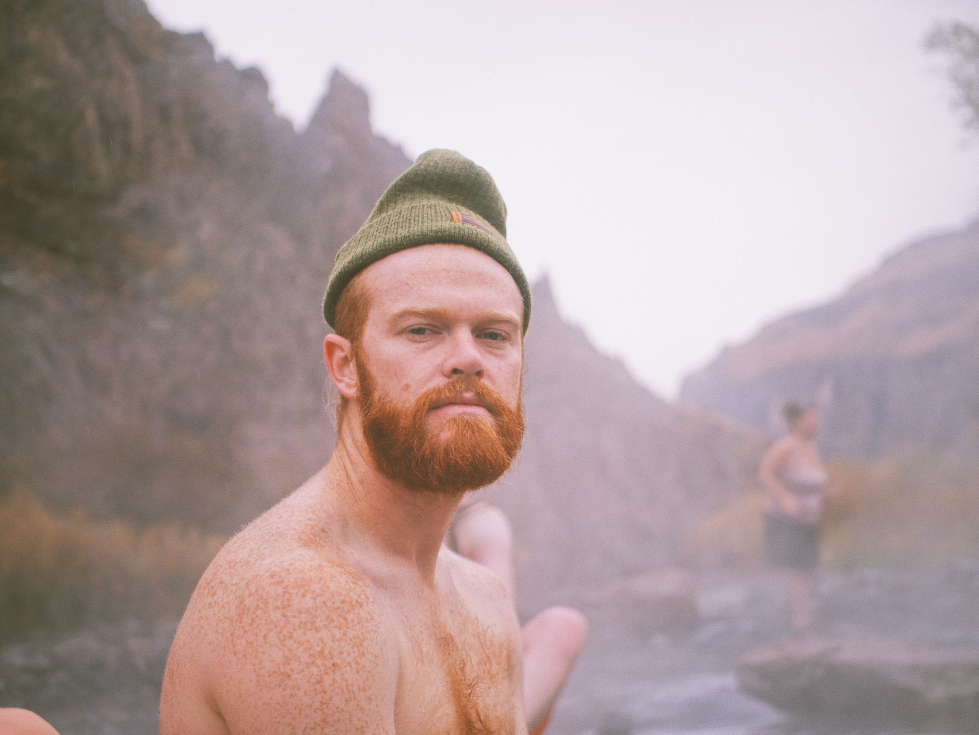 A shirtless man with a beard is wearing a beanie and looking at the camera.