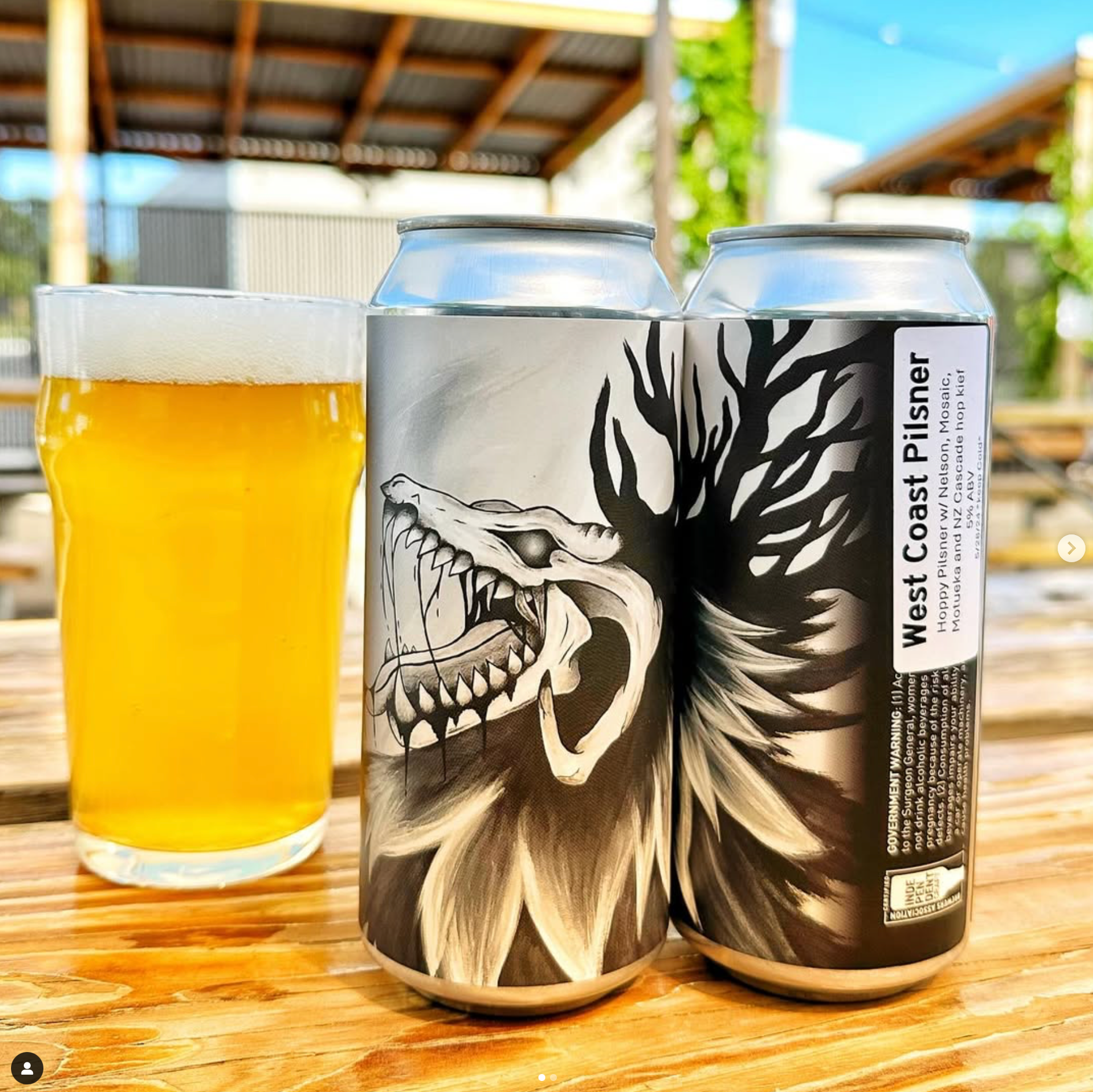 Two cans of west coast pilsner are sitting on a wooden table next to a glass of beer.
