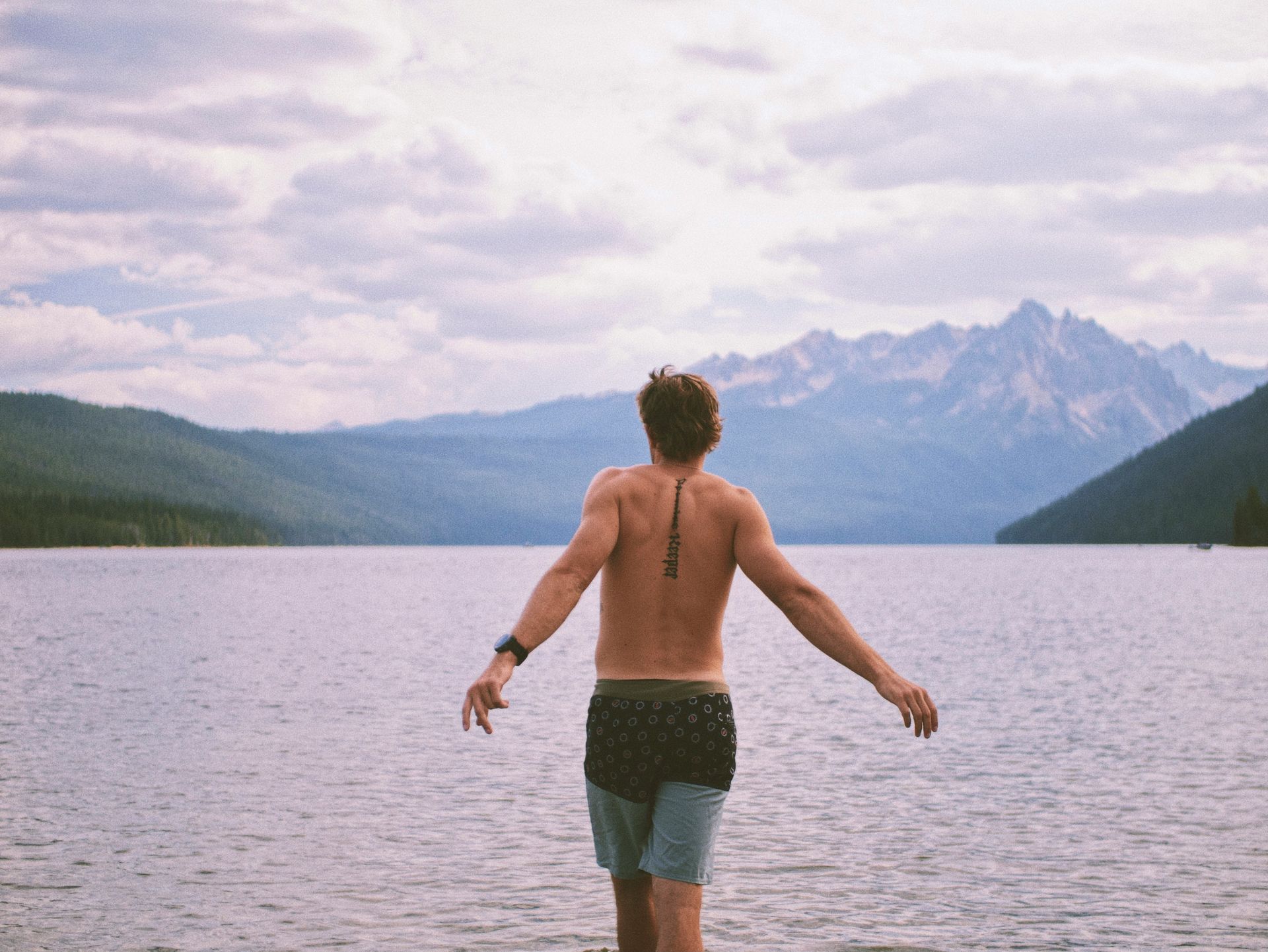 A shirtless man is walking into a lake with mountains in the background.