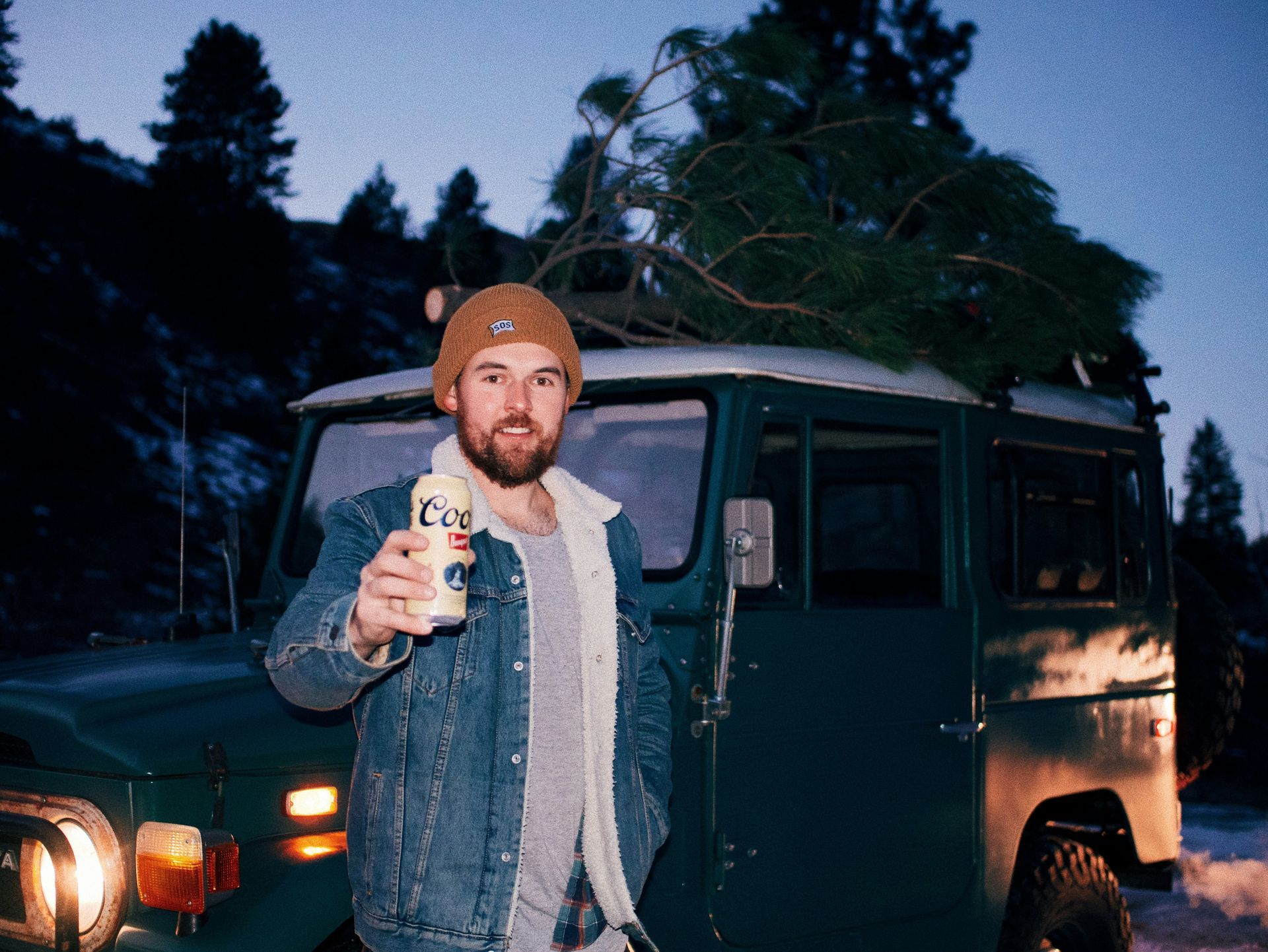 A man is standing in front of a jeep with a christmas tree on top of it.
