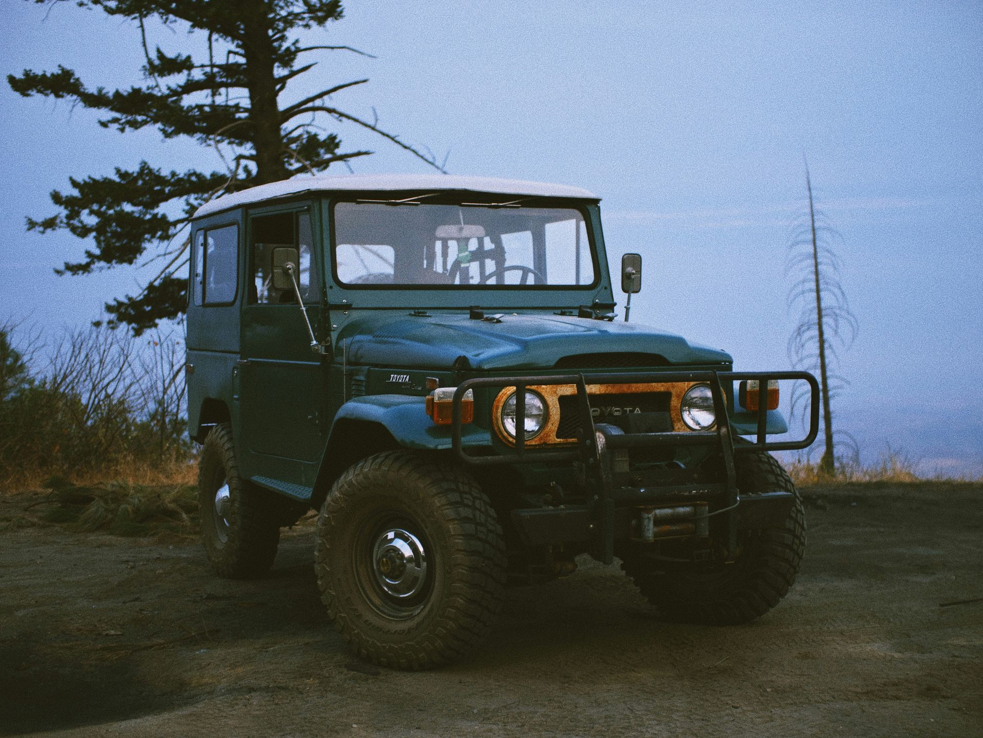 A blue jeep is parked on a dirt road next to a tree.