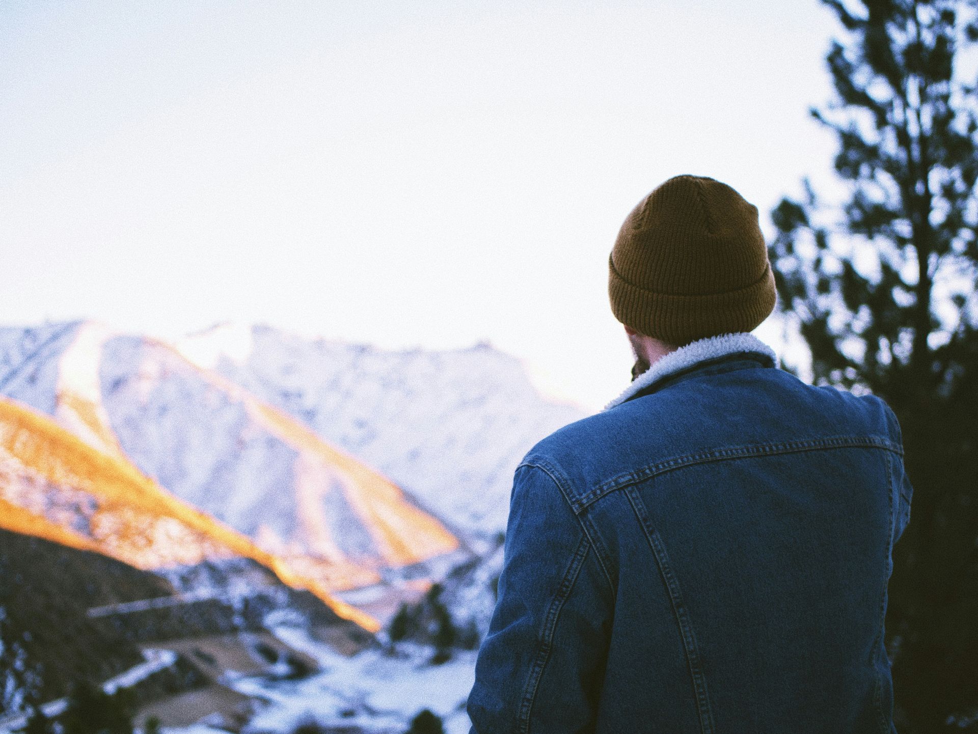 A man in a denim jacket is standing in front of a snowy mountain.