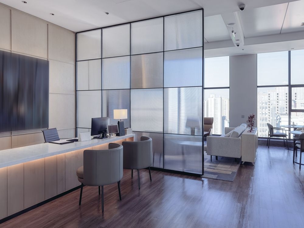 Modern office with glass wall separating desk area and seating. Includes computer, chairs, and large windows.