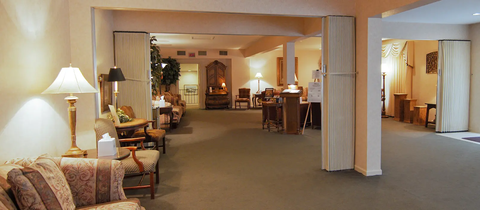 Interior of a funeral home with tan walls, carpet, and seating areas. Soft lighting and open spaces create a somber atmosphere.