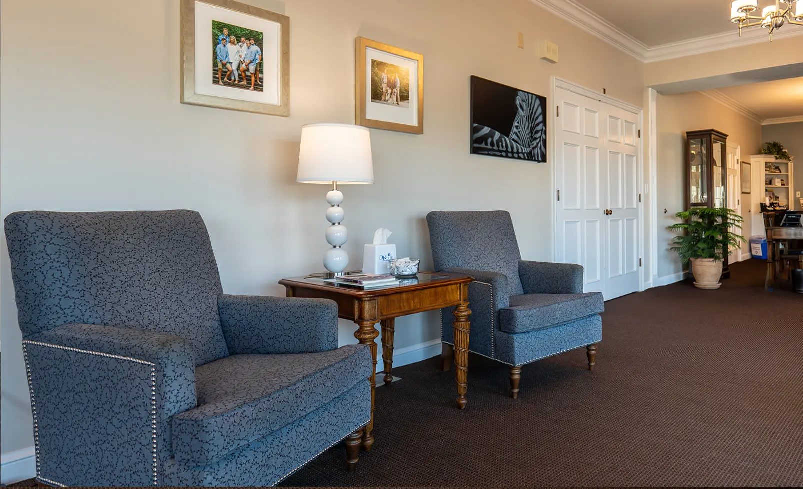 Two gray armchairs next to a side table with lamp and tissues, in a waiting area with artwork and a doorway.