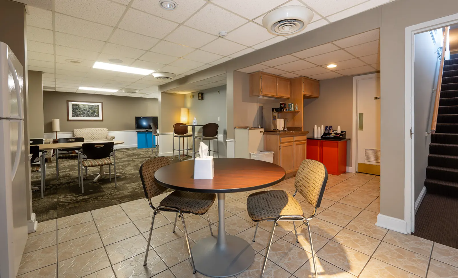 Communal room with round table, chairs, kitchenette, seating area, and stairs. Neutral tones.