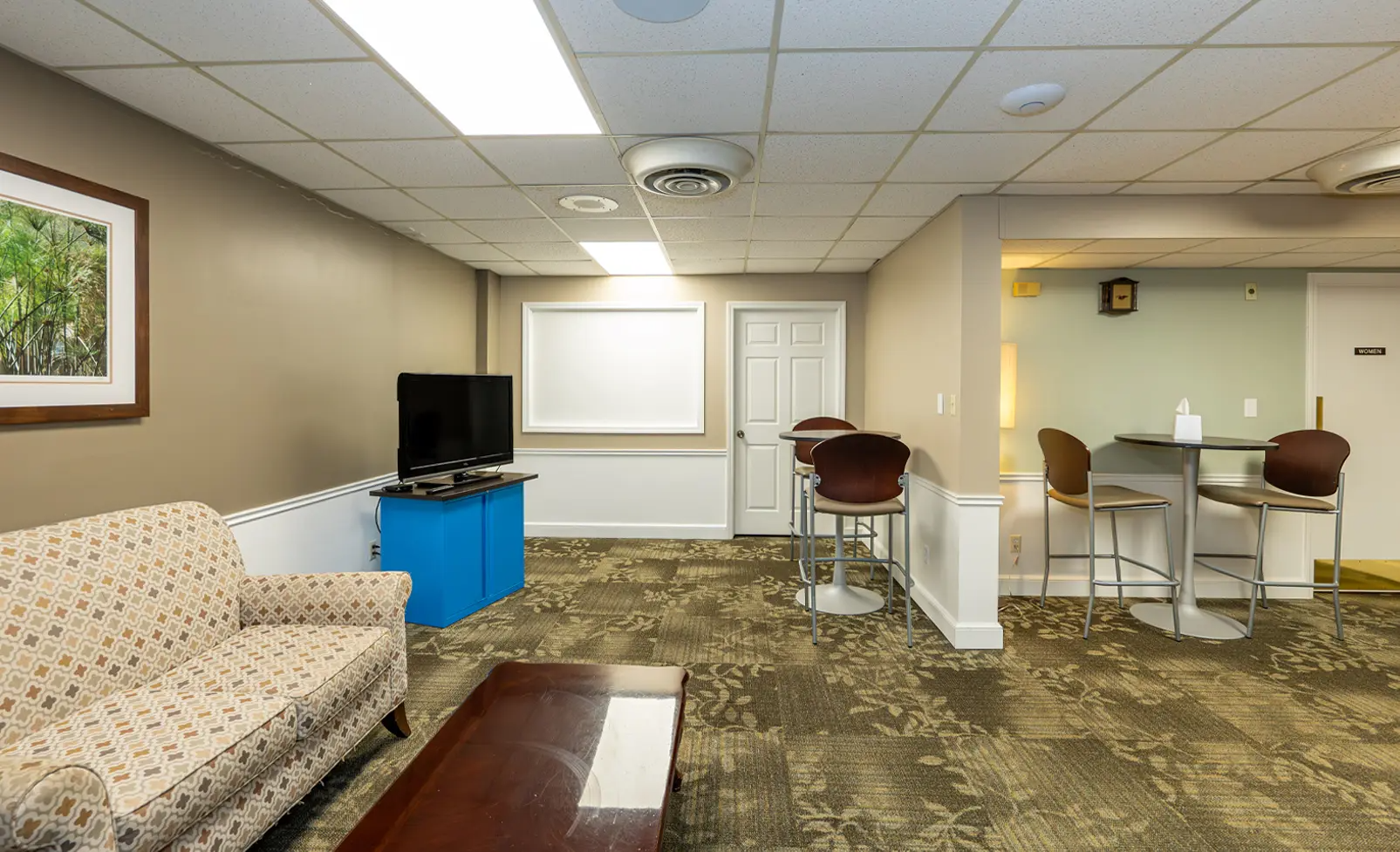 Lounge area: beige sofa, TV on blue stand, high-top tables with stools, door, and carpet.