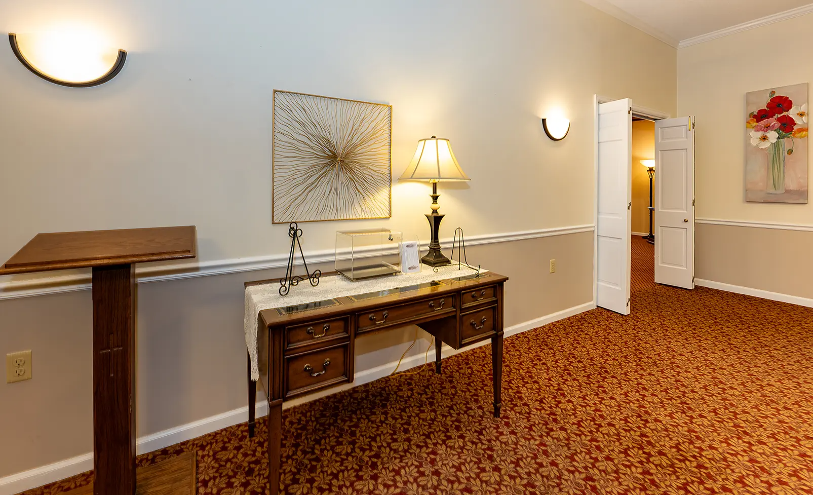 Wooden desk with lamp, artwork, and a podium in a room with floral carpet.