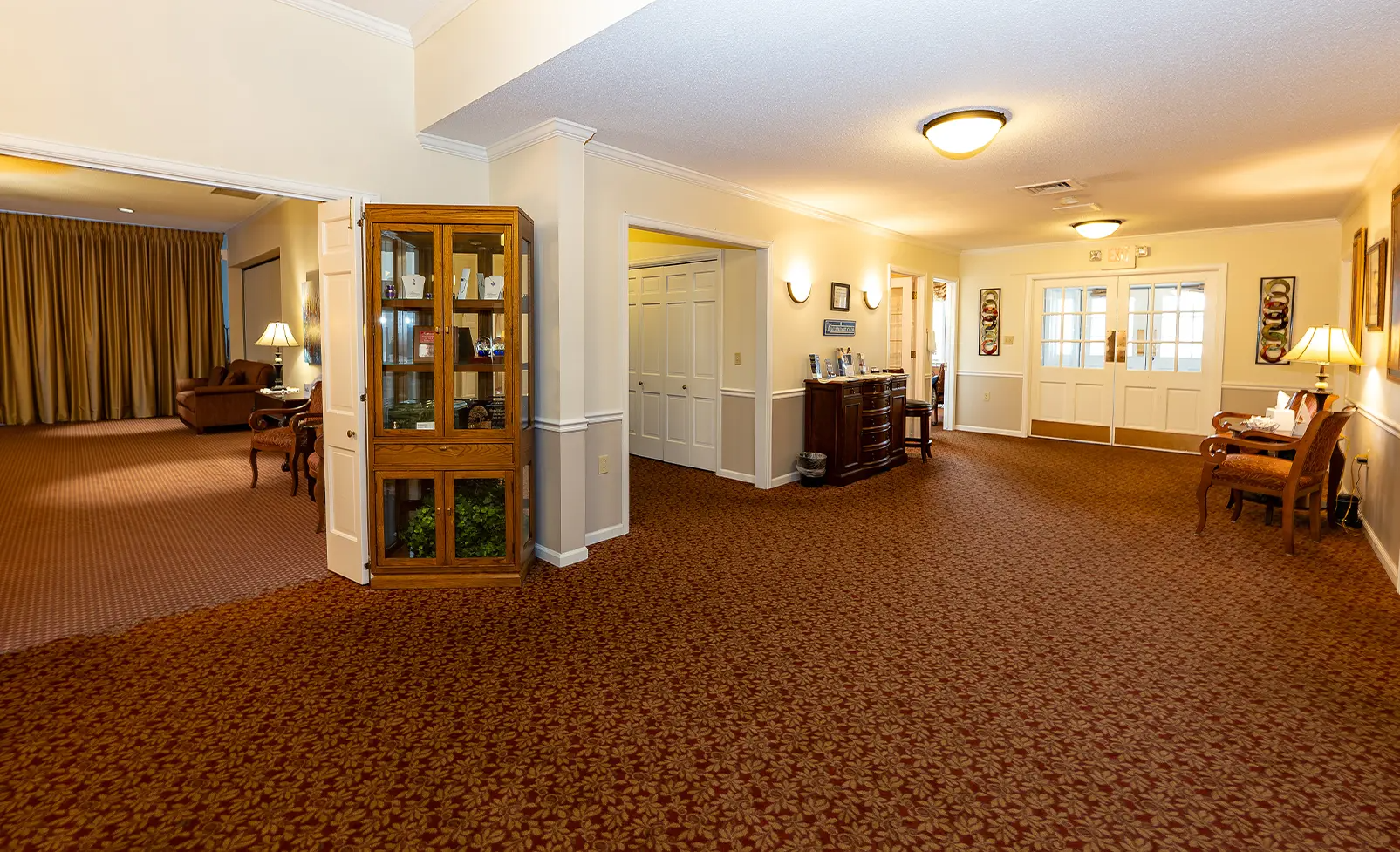 Large, carpeted hallway with glass cabinet, wooden chairs, and doors.