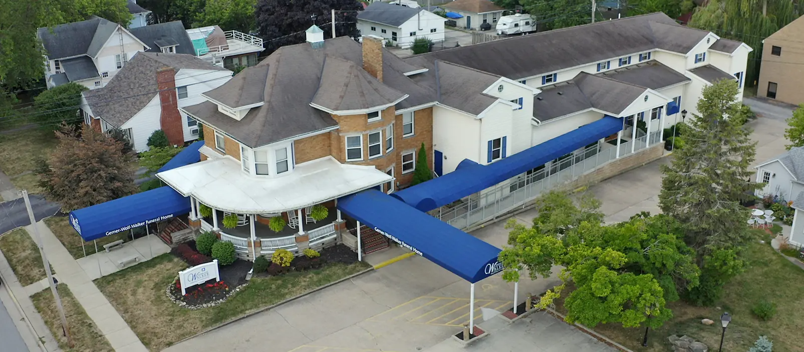Aerial view of a large building with a blue awning. There are trees and other buildings in the background.