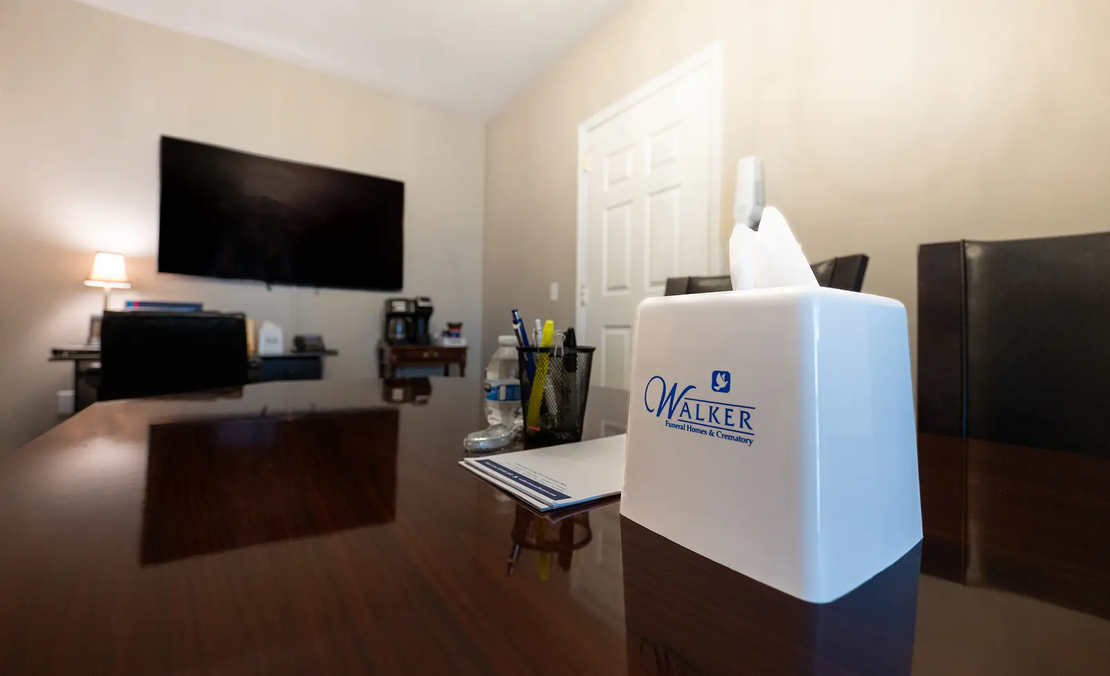 A wood desk with a tissue box in an office with a mounted TV and closed door.