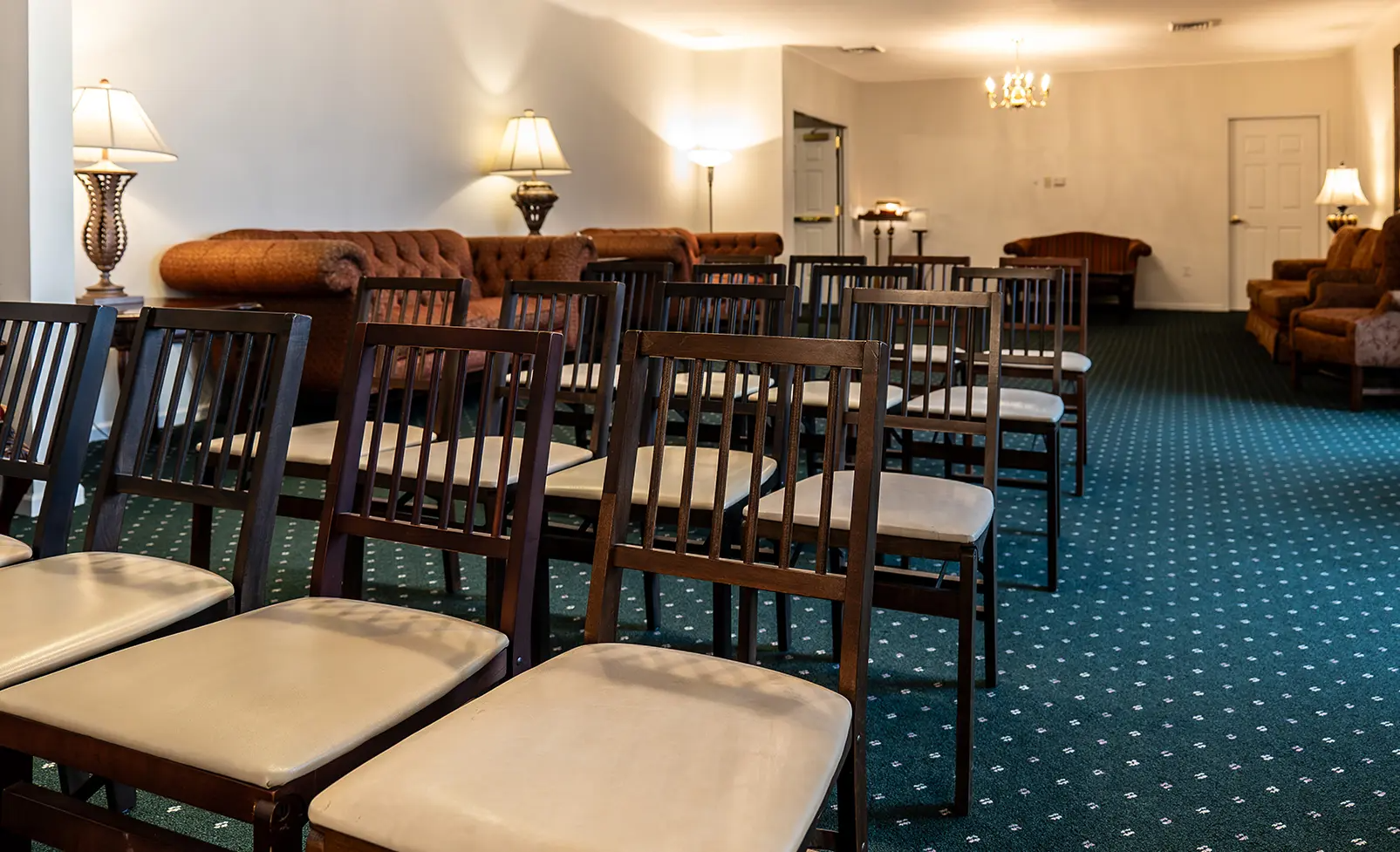 Rows of chairs face sofas in a room with lamps and a patterned green carpet.