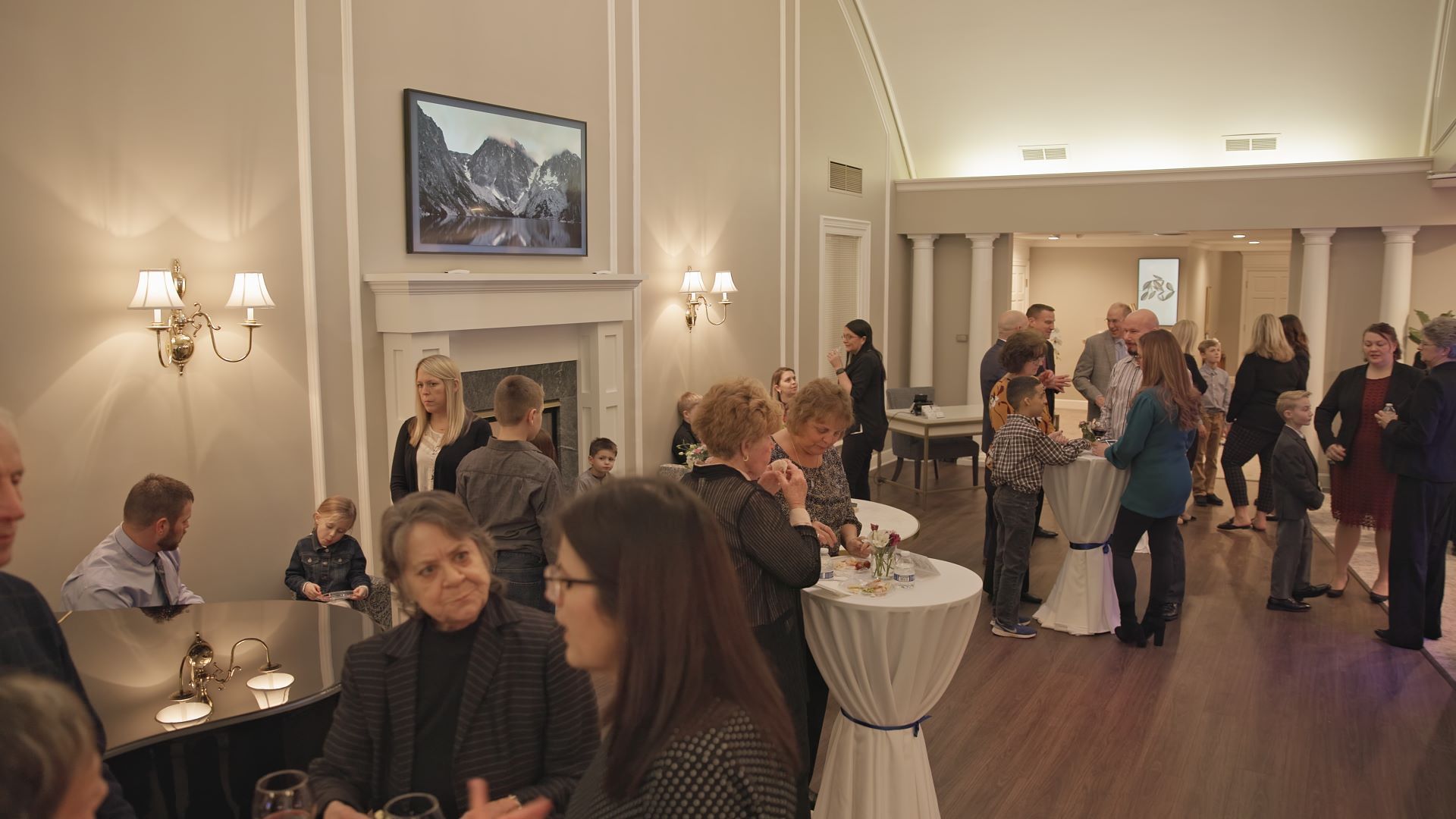 People mingling in a brightly lit event space with round tables, light walls, and a fireplace.