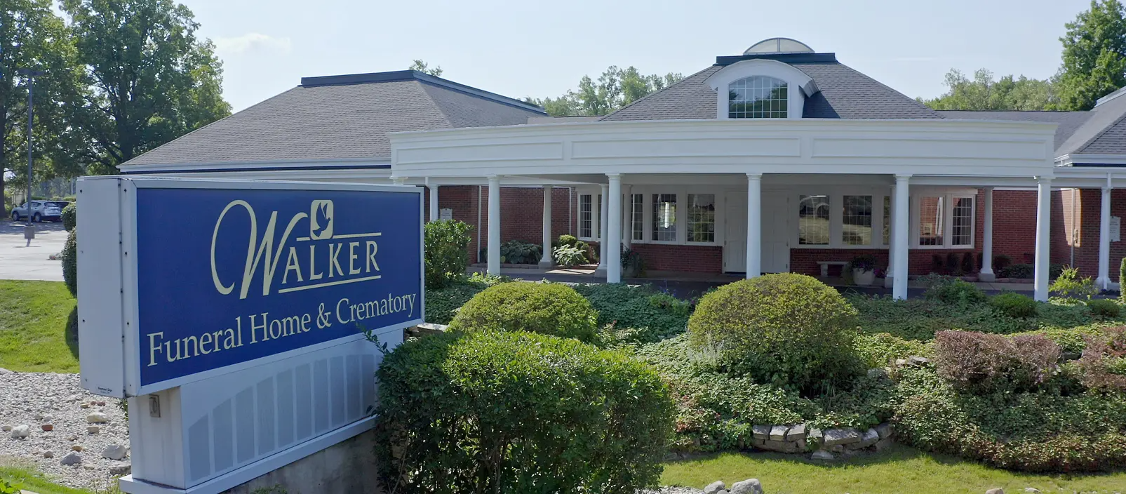 Walker Funeral Home & Cremation sign in front of building with white columns and a dark roof. Green bushes in the foreground.