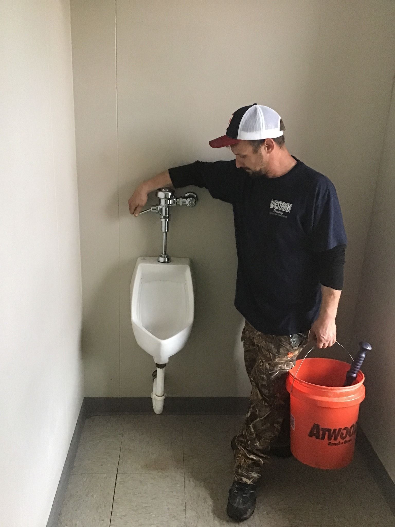 A man is standing next to a urinal in a bathroom holding an orange bucket.