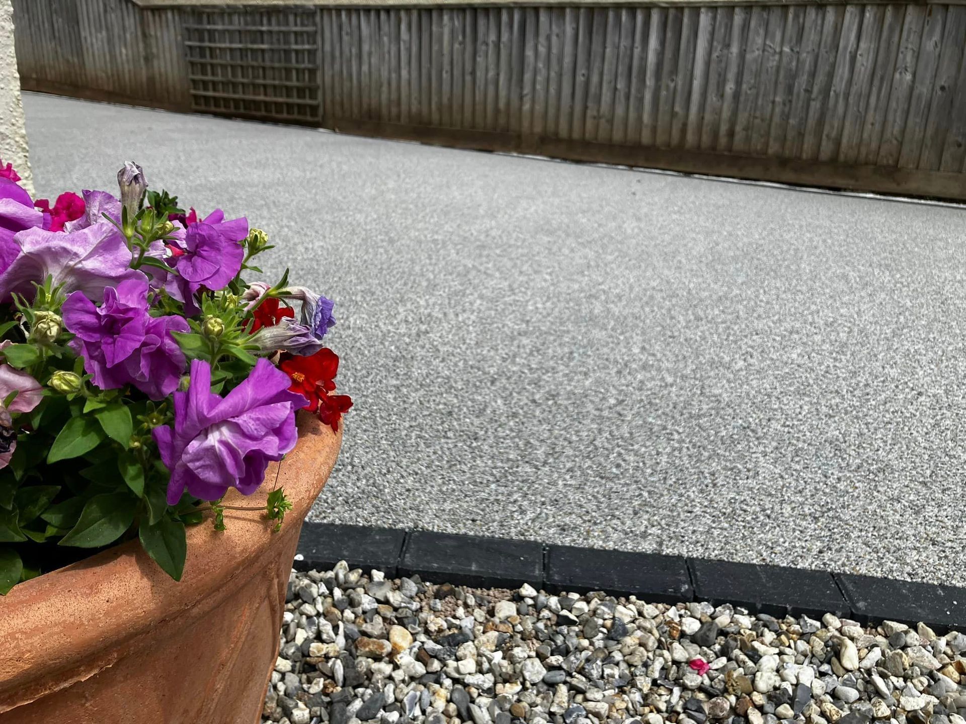 Grey resin driveway with a black border next to a flower pot with purple flowers.