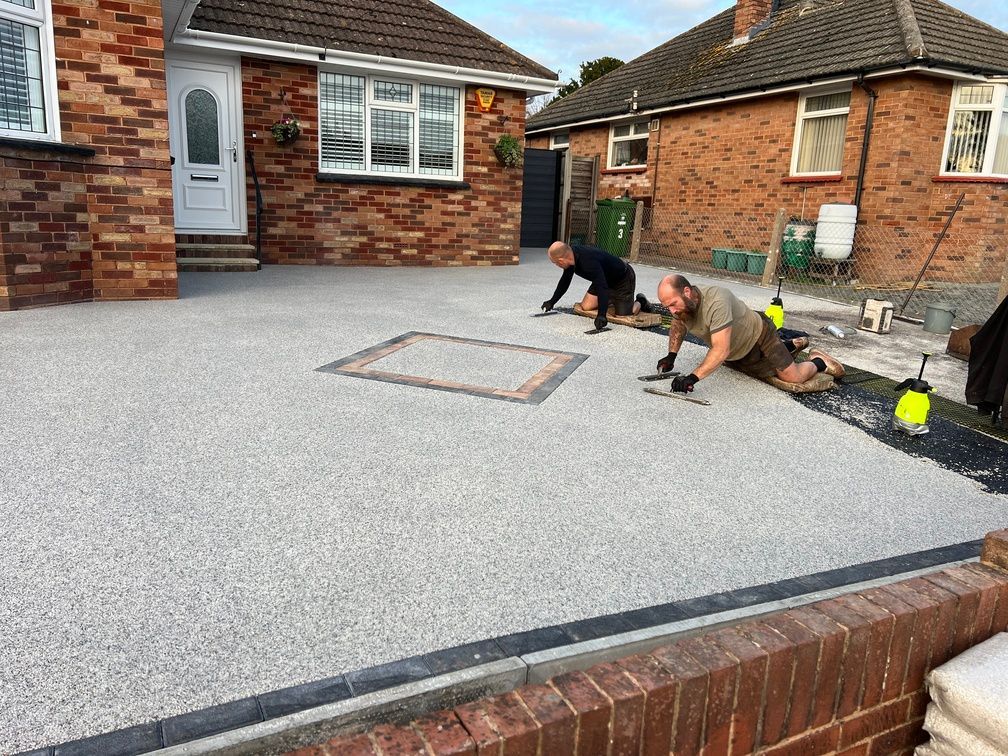 Grey resin driveway being installed by 2 men, outside a red brick house.