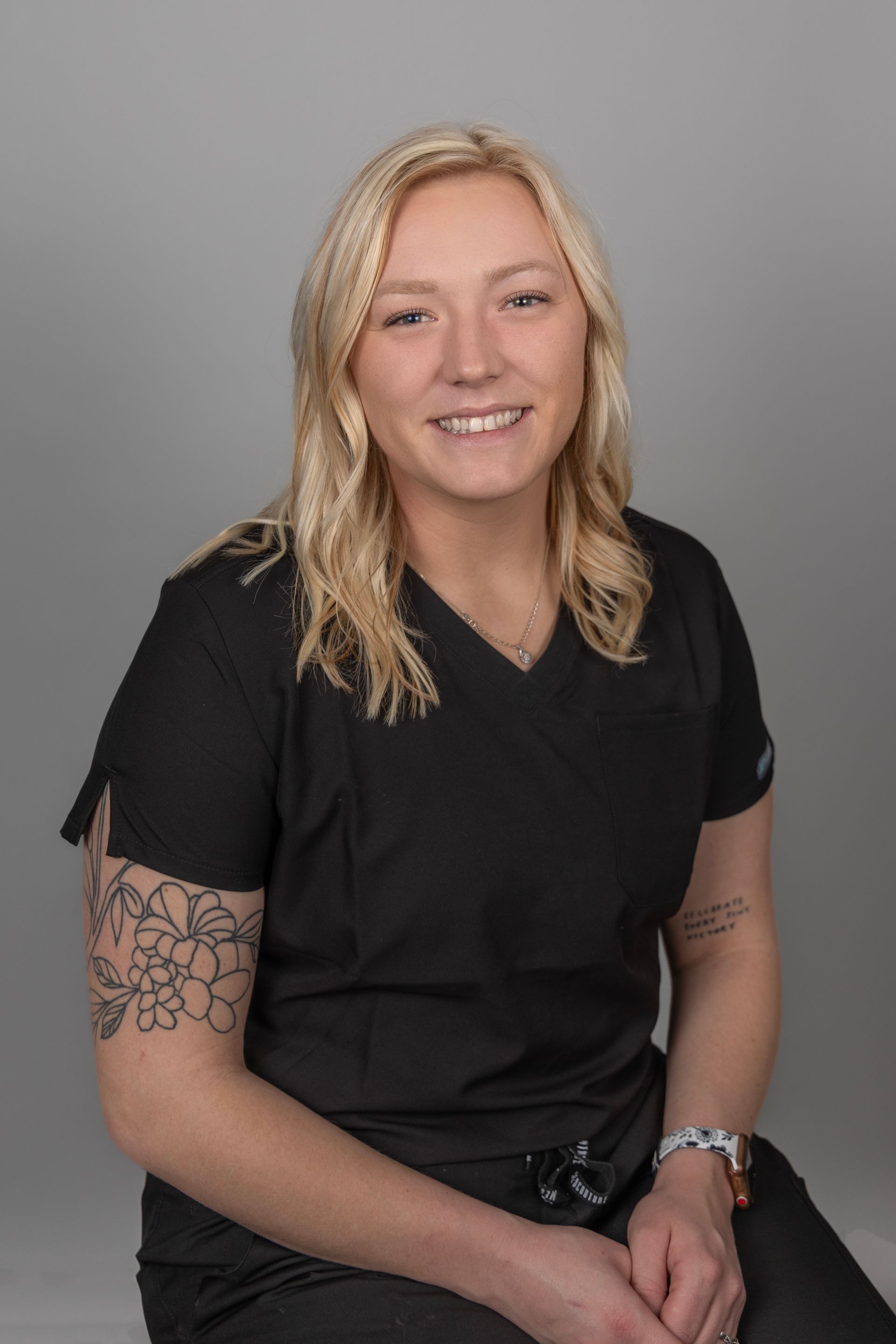 A woman in a black scrub top is sitting down and smiling.