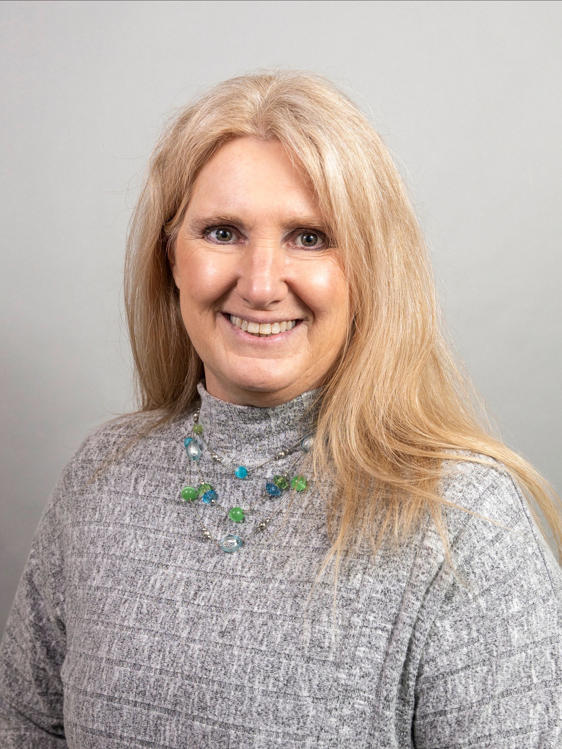 A woman wearing a grey sweater and a green necklace is smiling for the camera.