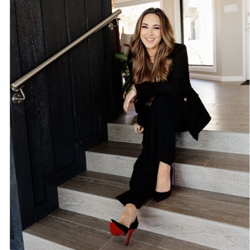 Woman in black suit and red heels, sitting on steps, smiling.