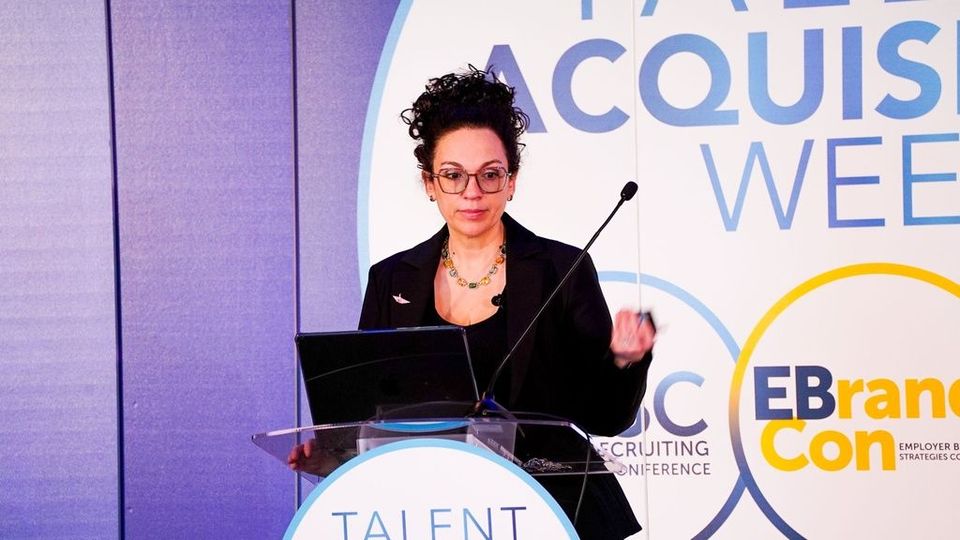 Woman at podium, presenting at conference; purple backdrop, logo, holding pen.