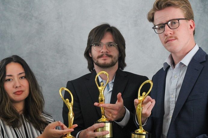 Three people holding gold trophies. They stand in front of a gray background. One person points to a trophy.
