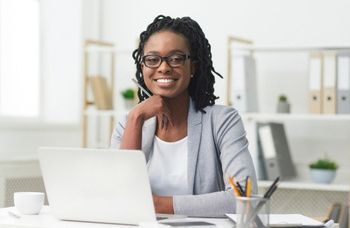 Woman in glasses at desk with laptop, smiling and holding her chin.