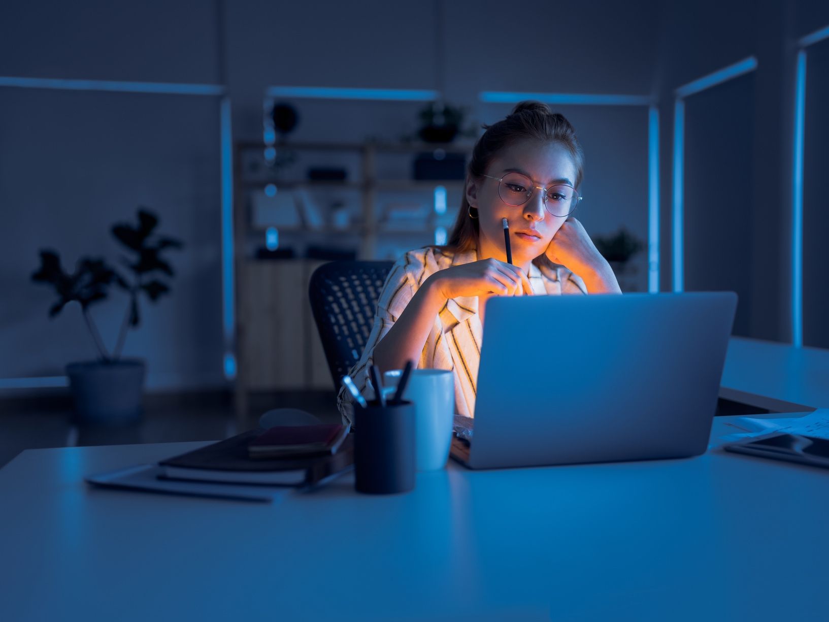 Woman working on a laptop in a dimly lit office at night. She has a pen to her lips.