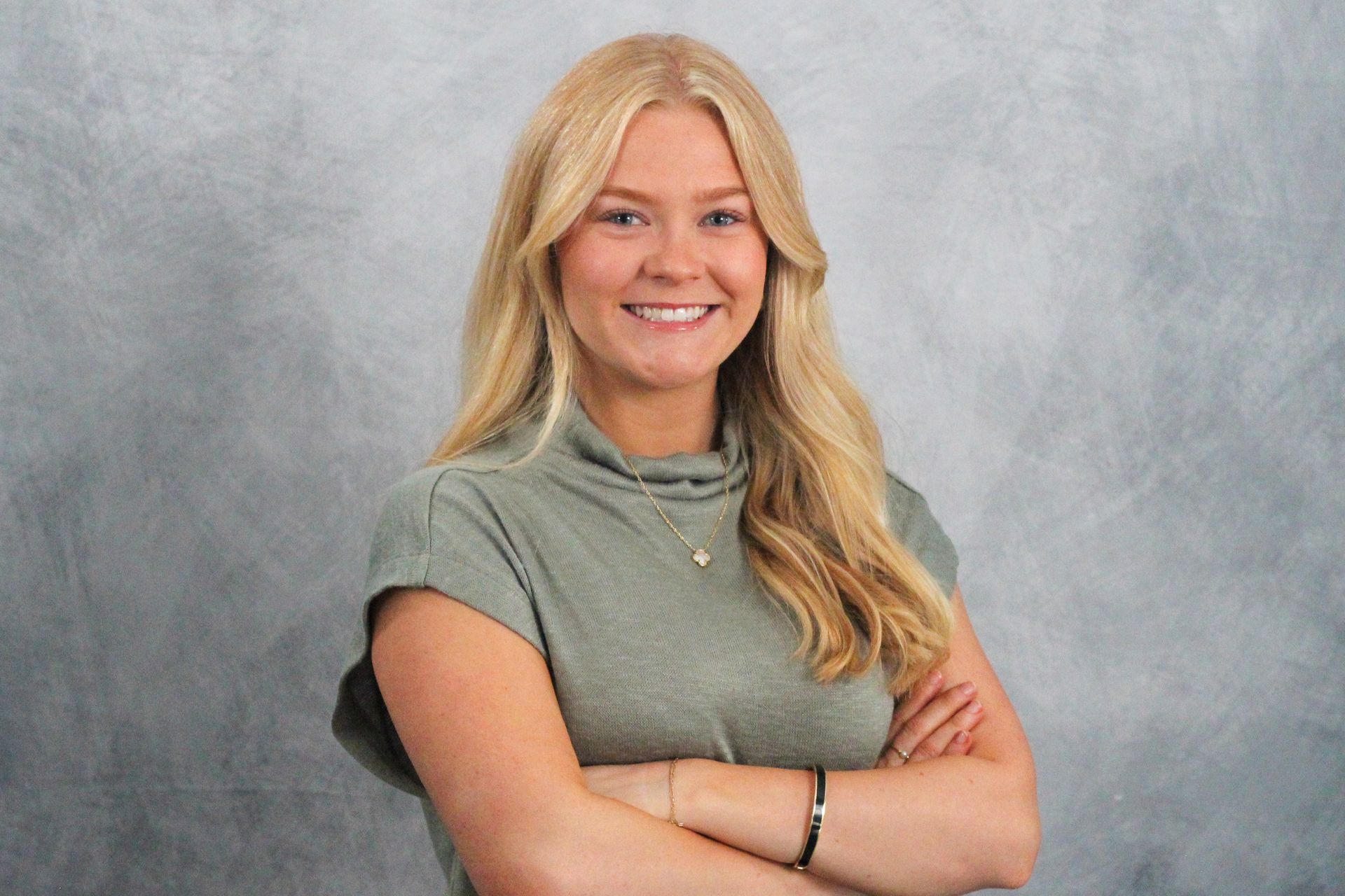 Blonde woman smiling, arms crossed, wearing a green turtleneck, against a gray background.