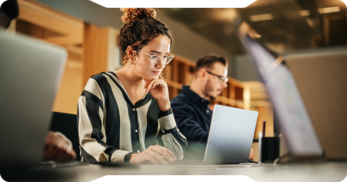 Woman with glasses working on a laptop in a well-lit office. A man is working in the background.