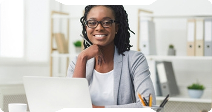 Woman in glasses smiles at her desk, working on a laptop.