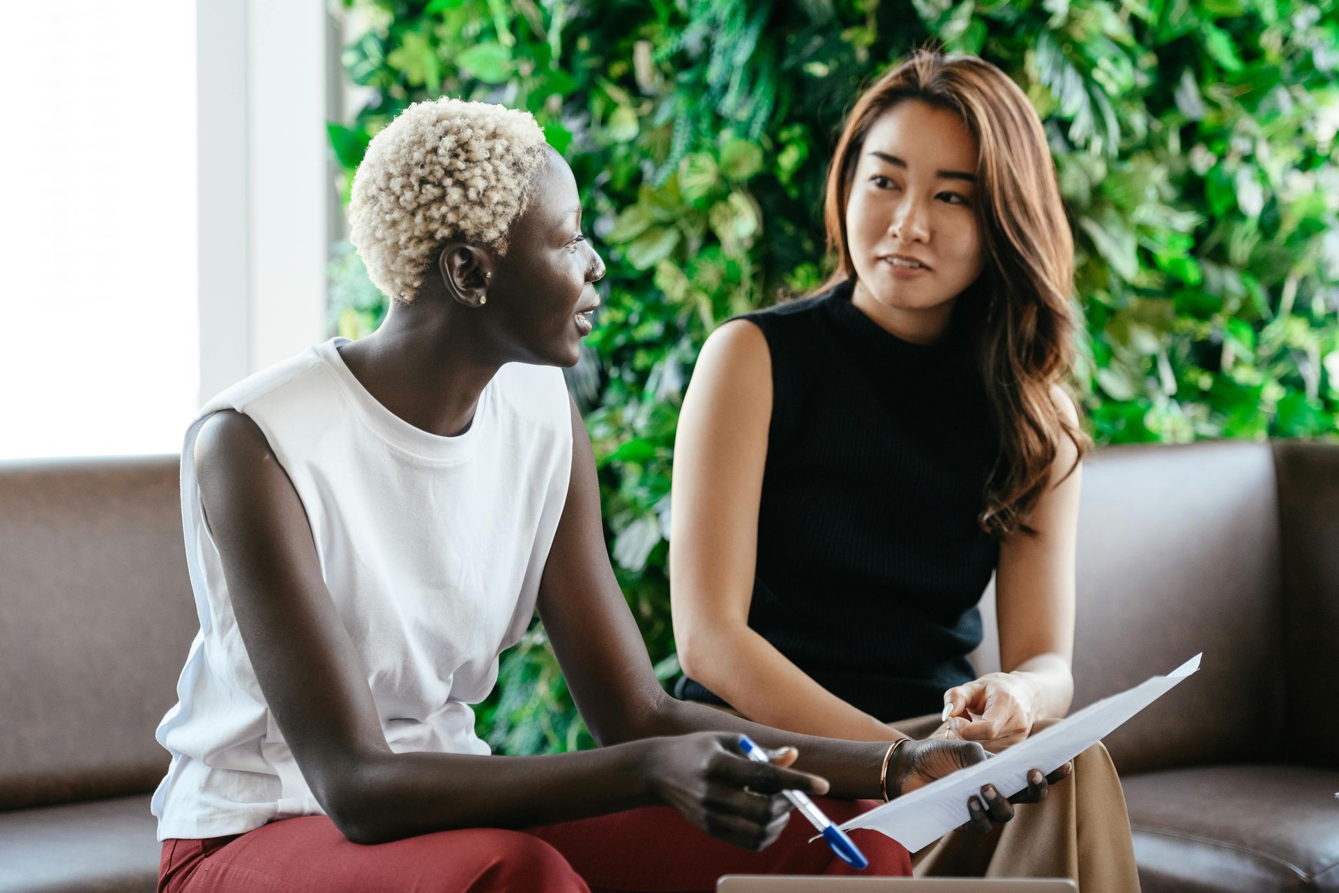 Two women in business attire reviewing documents on a couch, with greenery in the background.