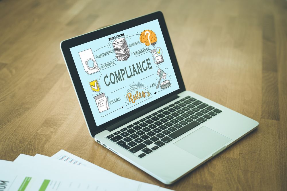 Laptop on a wooden desk showing a “Compliance” slide, with papers beside it.