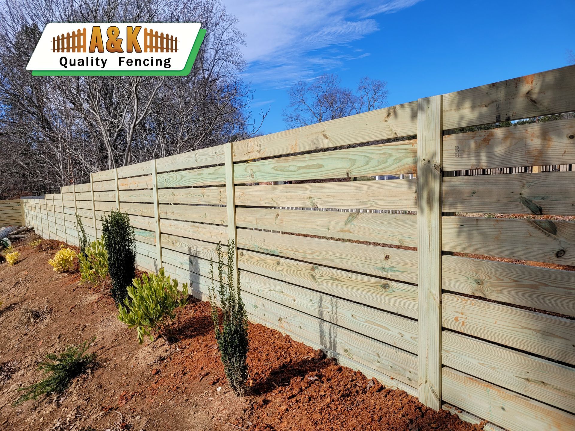 A wooden Horizontal fence is surrounded by plants and dirt in a yard.