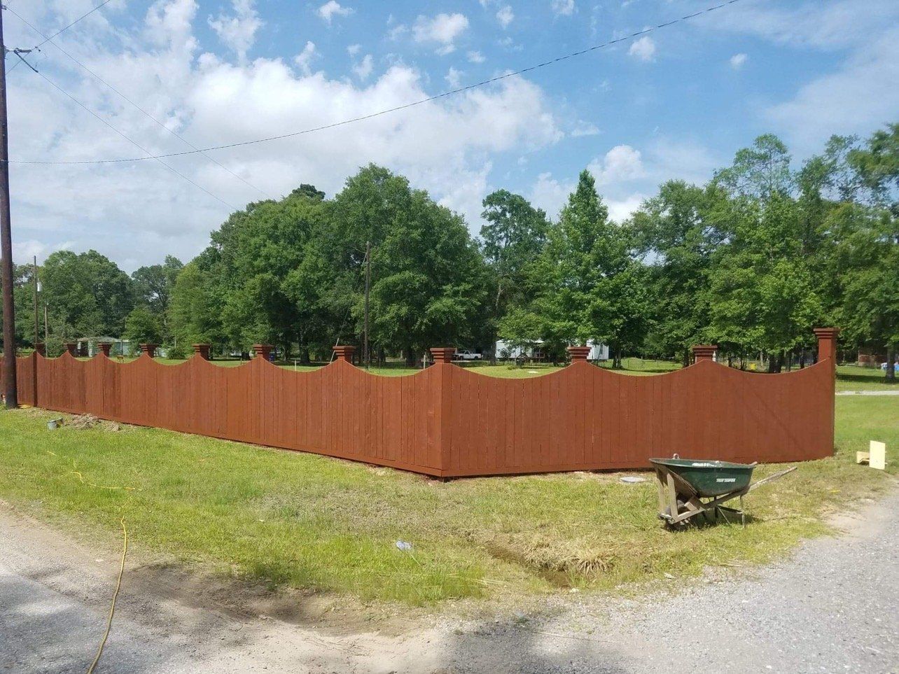 A wooden fence surrounds a grassy field with trees in the background and a wheelbarrow in the foreground.