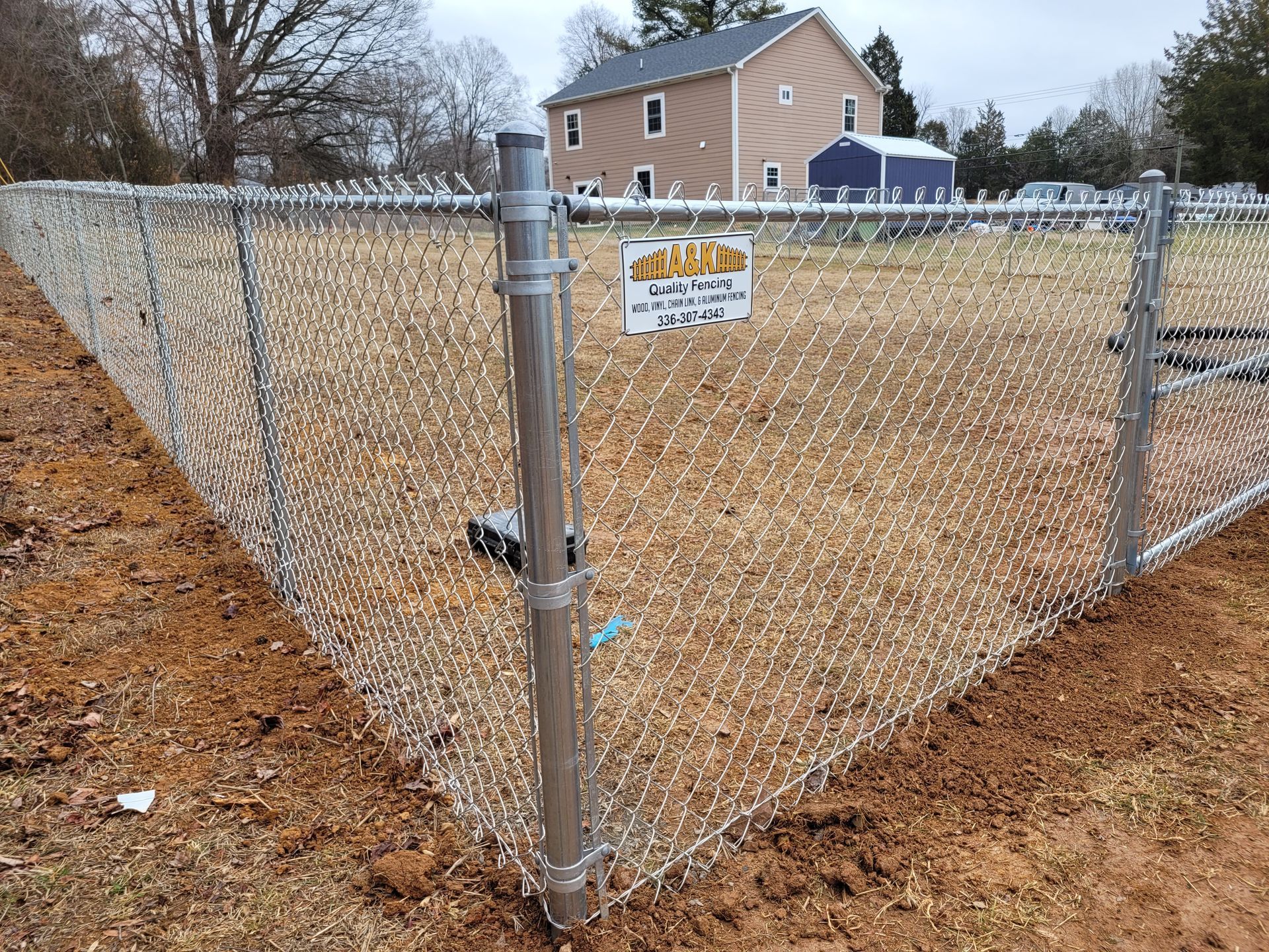 A chain link fence surrounds a dirt field with a house in the background.